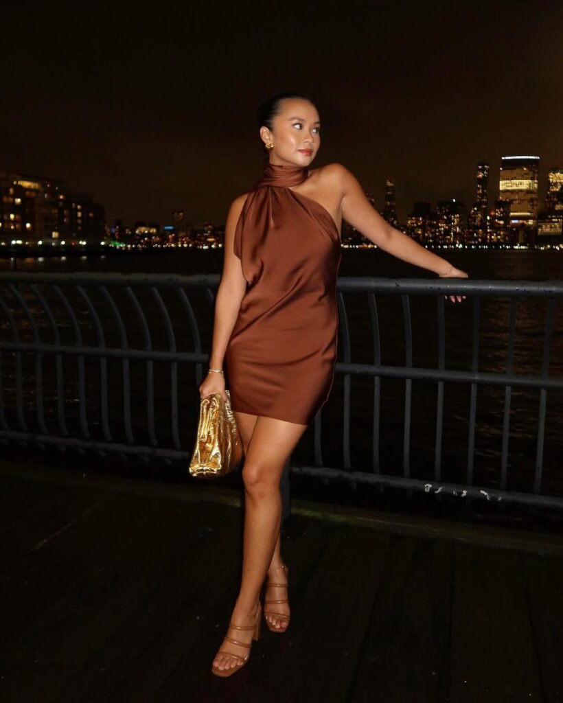 Woman in a one-shoulder satin bronze mini dress and nude heels by the water — a glamorous Night Out Outfit.