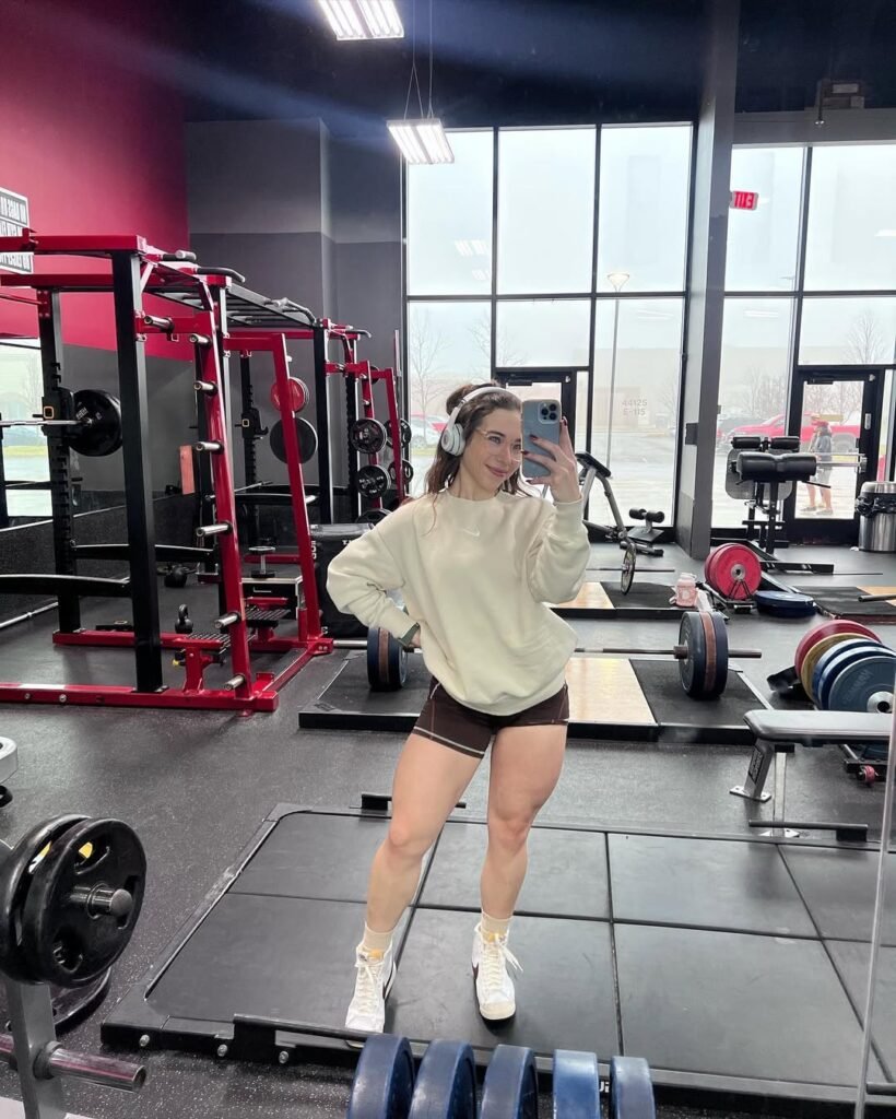 Sporty gym outfit inspo inside the gym, pairing a cream sweatshirt with brown shorts and white sneakers for a cozy lift-day fit.