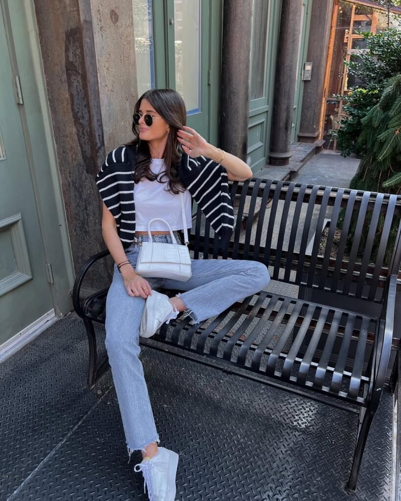  Woman sitting on a black bench in a striped sweater, white top, and jeans – perfect for casual outfit inspo.