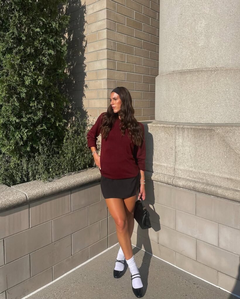 Fall-ready outfit inspo with a burgundy sweater and black mini skirt styled with socks and flats.