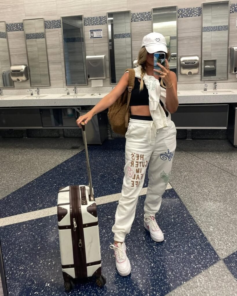 Woman in stylish joggers and crop top posing with luggage in an airport bathroom – a comfy and trendy airport outfit.

