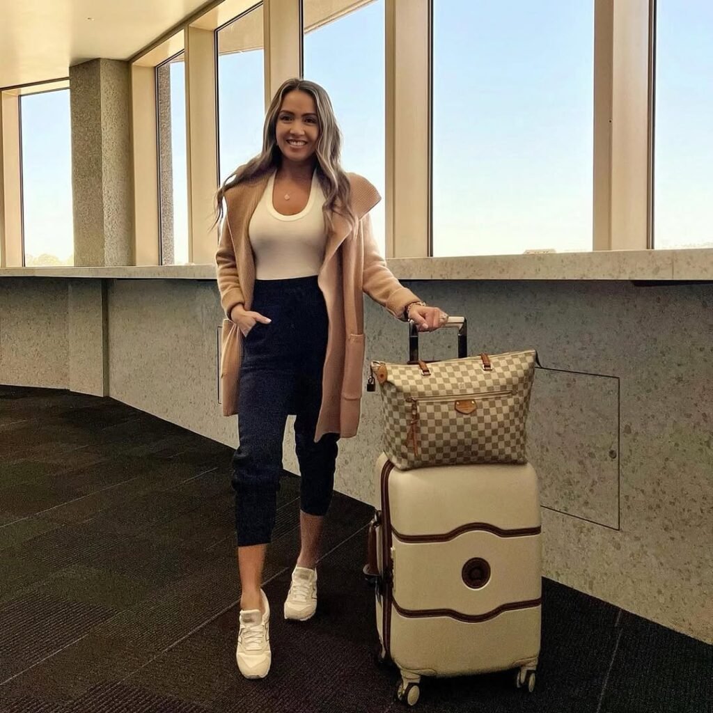 Woman in a white tank, camel coat, and sneakers with checkered bag – cozy and polished airport outfit style.