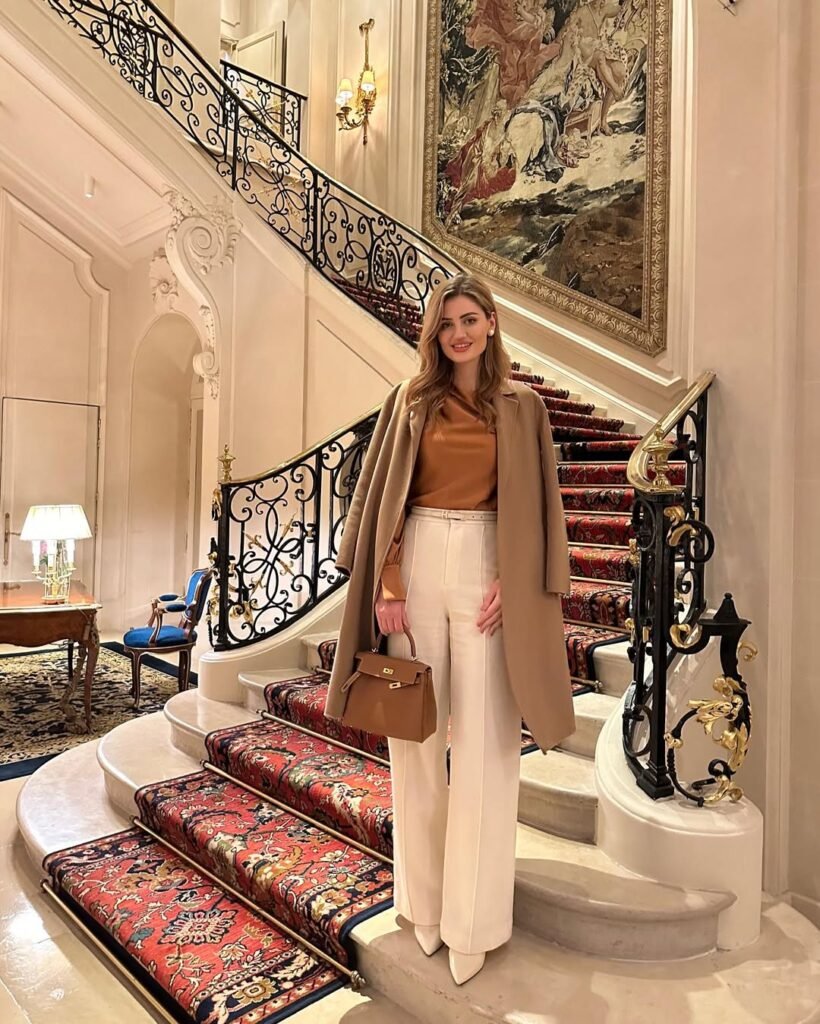 Woman on a grand staircase in a caramel blouse and cream trousers paired with a tan overcoat – timeless elegant outfit for luxury settings.