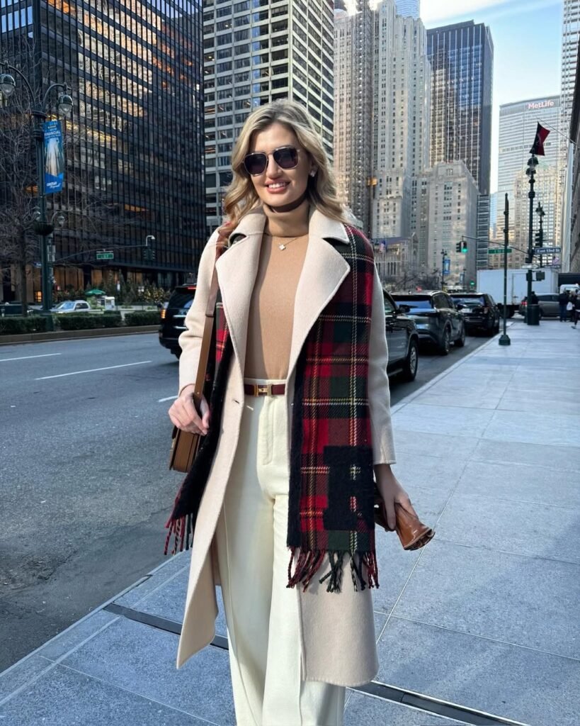 Classy woman in a long cream coat, tan top, white trousers, and a tartan scarf – elegant outfit for winter city fashion.