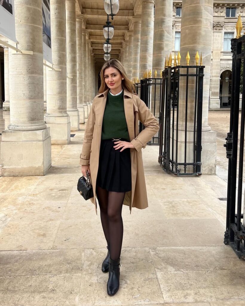 Sophisticated green sweater, pleated black skirt, and trench coat – elegant outfit for a day out.