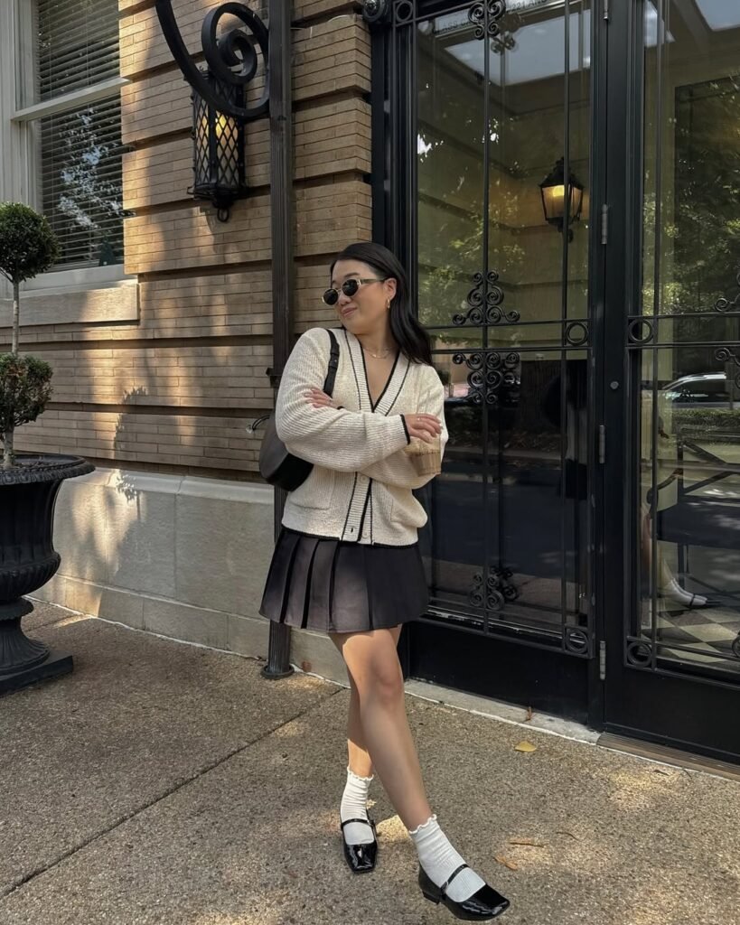 Chic cardigan outfit featuring a cream button-up cardigan paired with a black pleated mini skirt, white socks, and black Mary Jane shoes.