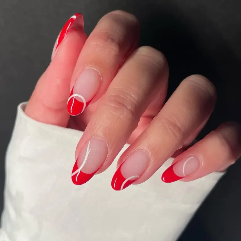 Close-up of a hand with long almond-shaped nails featuring red tips with delicate white swirl accents, showcasing elegant Red Nail Ideas.