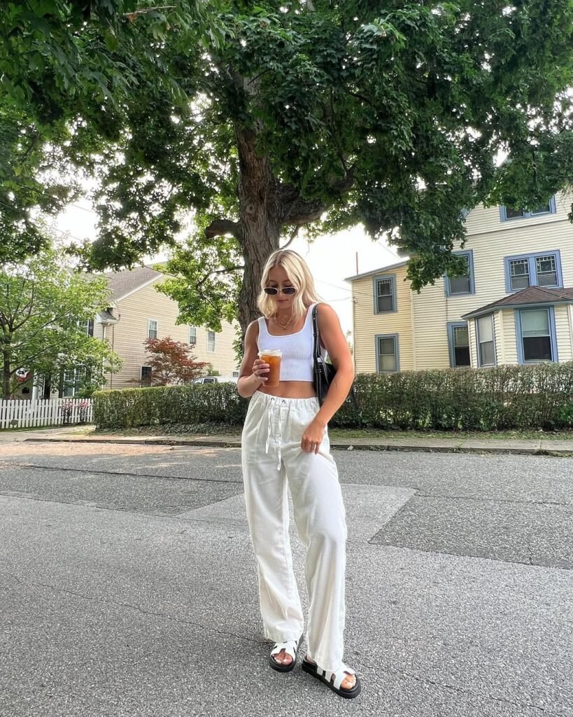 Woman walking outdoors wearing white linen pants paired with a white crop top and black sandals, holding an iced coffee.