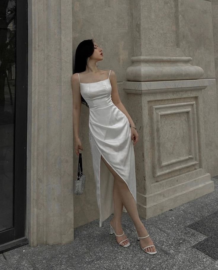 A woman wearing a satin white dress with spaghetti straps, striking a pose with a side slit, and elegant sandals, standing next to a beige building.