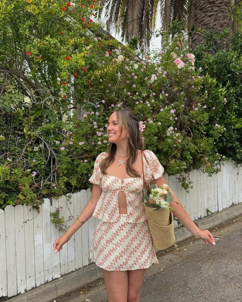  A woman in a light beige brunch outfit with a floral pattern, wearing a short skirt and top with a cut-out detail, holding a bouquet of flowers, smiling near a green backdrop of plants.