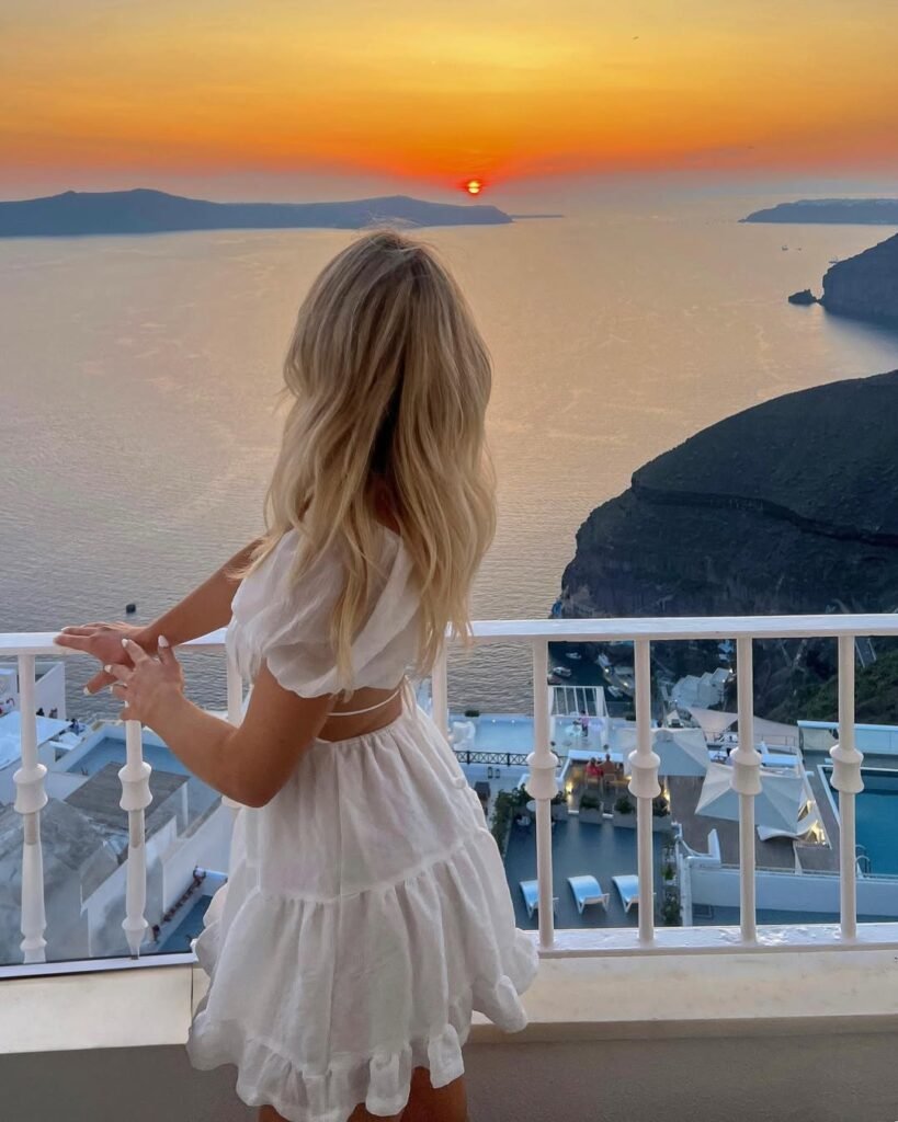 A woman in a white ruffled dress looking out at a stunning sunset over the sea from a balcony in Santorini, showcasing a romantic European summer outfit.