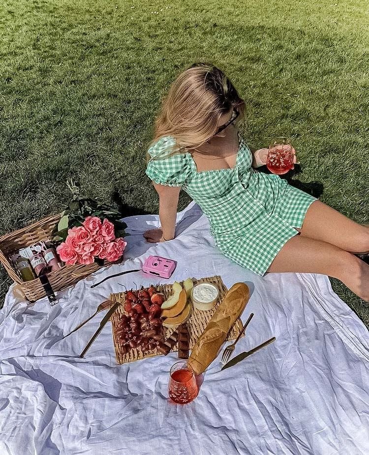 Picnic Date Outfits: Woman in a green gingham dress enjoying a picnic with flowers, fruit, and drinks on a sunny lawn.