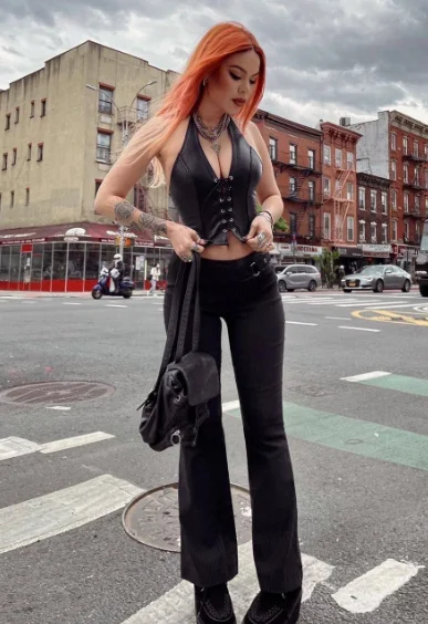 Grunge Outfits: Person with orange hair wearing a black halter crop top with lace-up detail, black high-waisted pants, and black boots standing on a street corner.