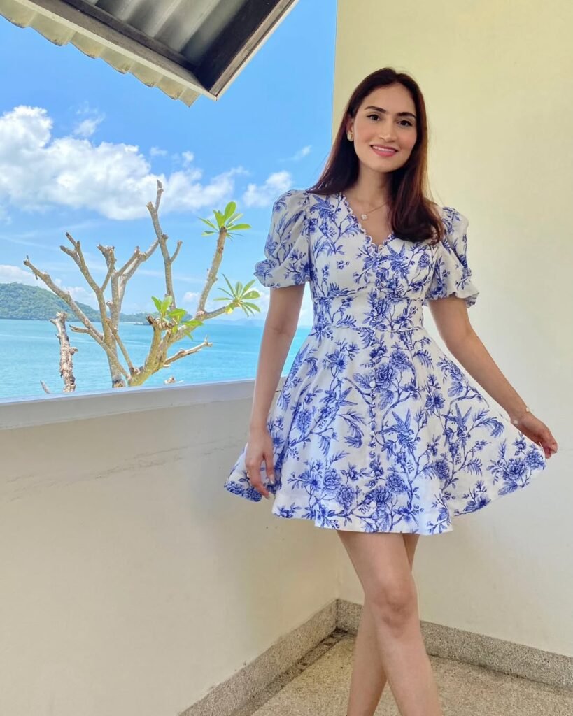 Woman in a blue and white floral mini dress with puff sleeves, smiling and posing near a balcony with a scenic ocean view, perfect for stylish outfits verano.