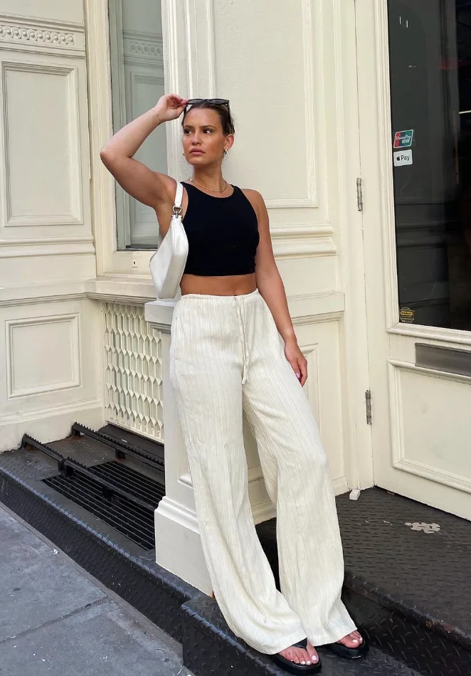 Woman wearing relaxed beige linen pants paired with a white cropped t-shirt, standing indoors near a window.