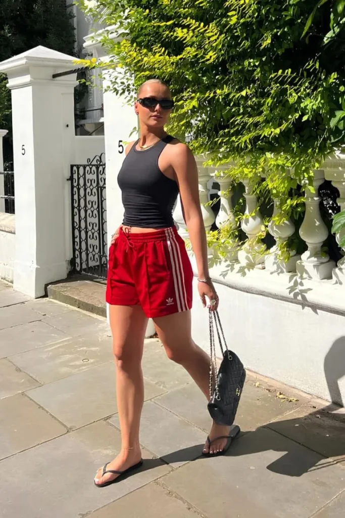 Sporty and edgy look with a black tank top and red Adidas shorts, accessorized with flip-flops and a shoulder bag — bold and fun outfit ideas summer.