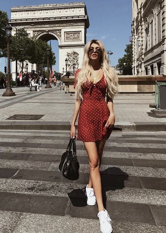 A woman crossing a street in Paris in front of the Arc de Triomphe, dressed in a red polka dot mini dress and white sneakers — a quintessential European summer outfit.