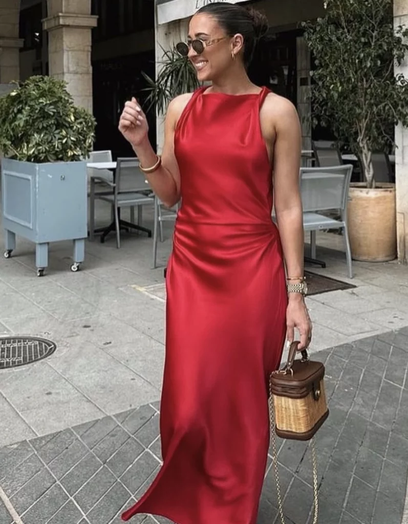A woman in a satin red dress with a sleek silhouette, accessorized with a basket-style handbag, walking outdoors with a confident smile.