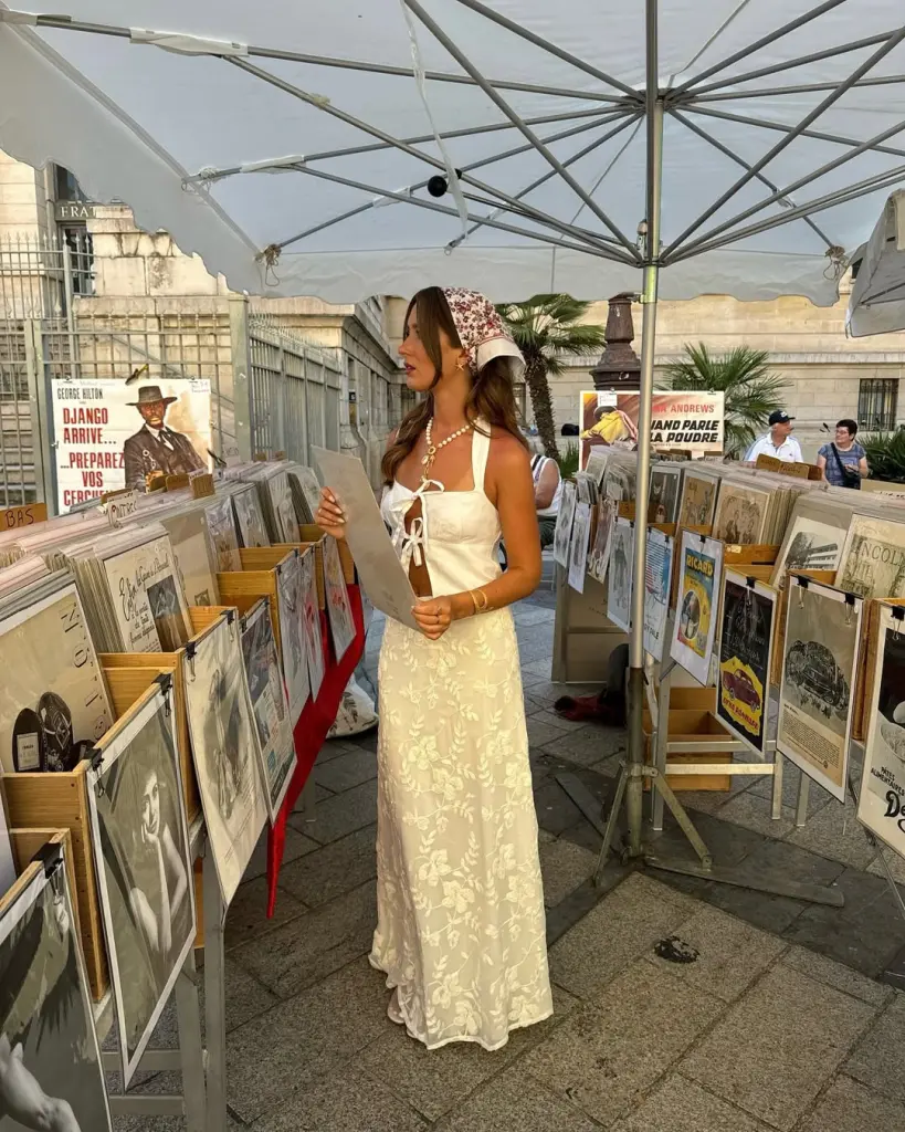 A woman browsing vintage posters in a white embroidered maxi dress and a floral headscarf under a market tent — European summer style.