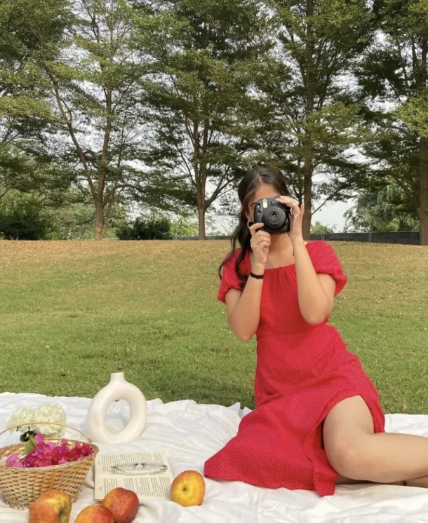 Picnic Date Outfits: Woman in a red dress taking photos with a camera at a cozy picnic set with books, apples, and flowers.