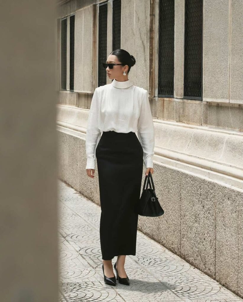 Stylish work attire featuring a white high-neck blouse tucked into a long black pencil skirt, completed with classic pumps and a structured black handbag for a refined and powerful ensemble.