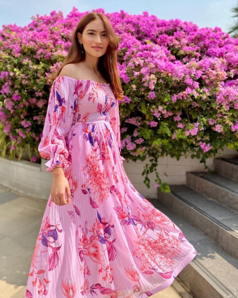 Young woman in a flowing pink floral off-shoulder maxi dress posing outdoors in front of vibrant pink flowers, showcasing a stylish and breezy summer outfit — Outfits verano.