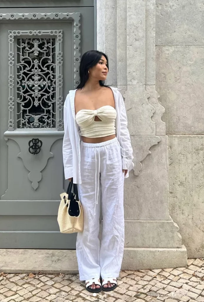 Woman in white linen pants with a cream crochet crop top and open linen shirt, holding a woven bag, standing against a textured wall. Stylish summer linen pant outfit.