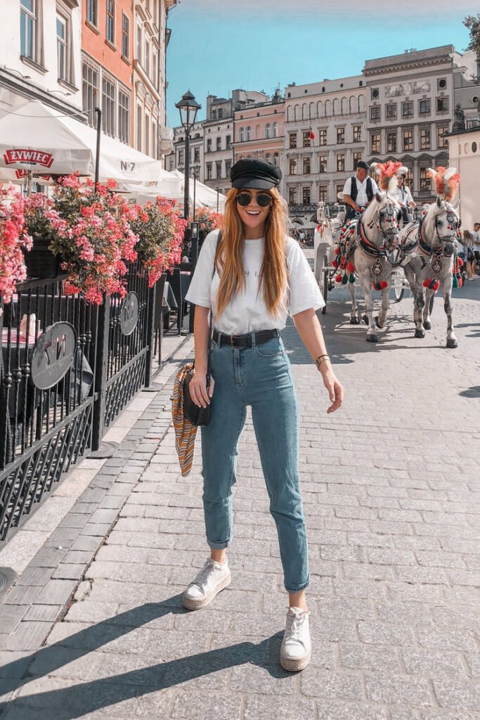 A woman in blue jeans and a white t-shirt, accessorized with a newsboy cap and round sunglasses, standing in a European square with horse-drawn carriages — a trendy European summer outfit.