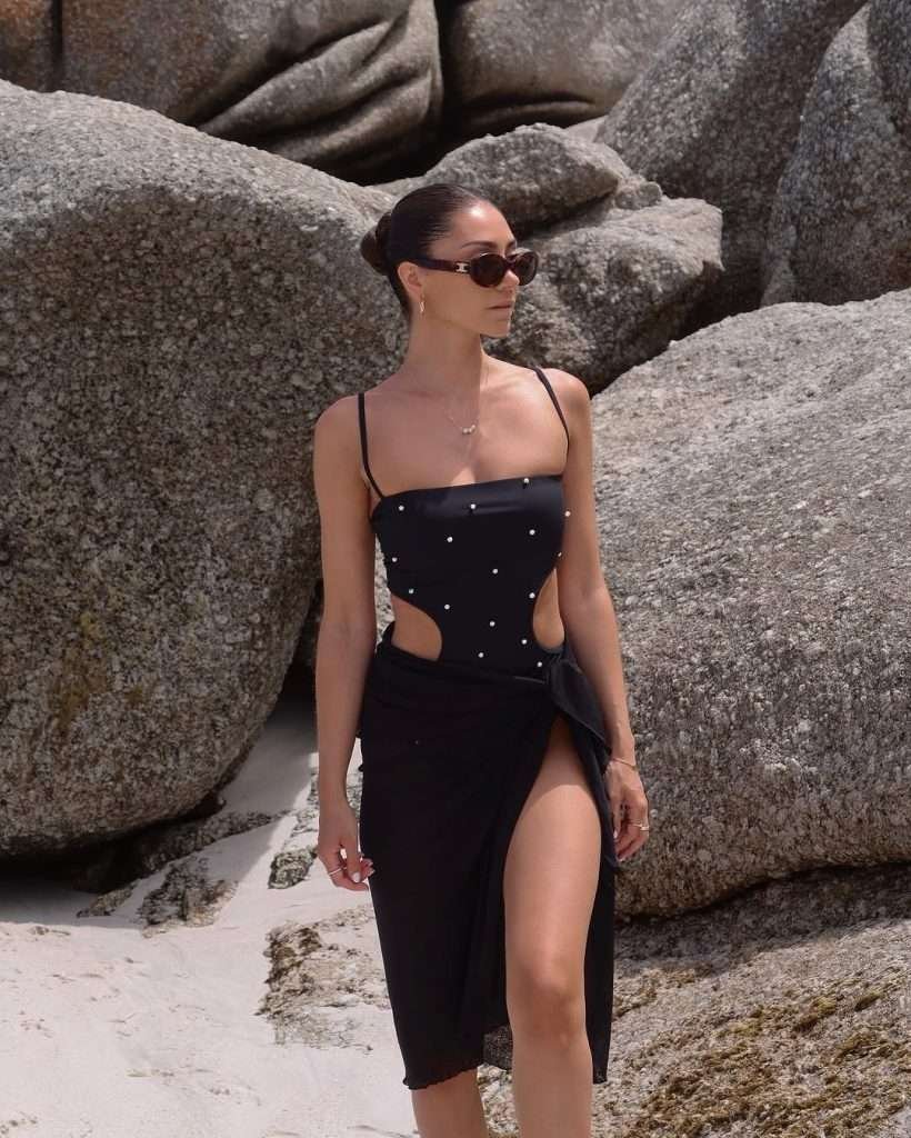 Beach outfit with a sleek black cut-out one-piece swimsuit adorned with pearls, styled with a matching black sarong and dark sunglasses, walking along a rocky beach.