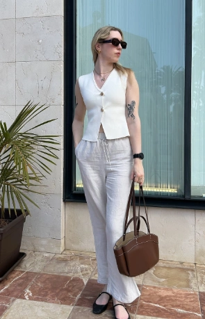 A woman wearing a sleeveless white buttoned vest with light linen pants and black flats, carrying a brown handbag; minimalist and elegant summer outfit idea.