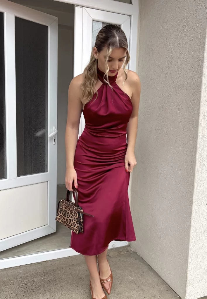 A woman wearing a satin burgundy dress with a halter neckline, holding a small leopard print handbag and looking down while standing near a doorway.