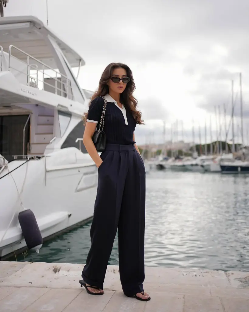 A woman standing by a marina in a navy outfit with a collared top and wide-leg trousers, accessorized with black heels and a quilted handbag — European summer style.