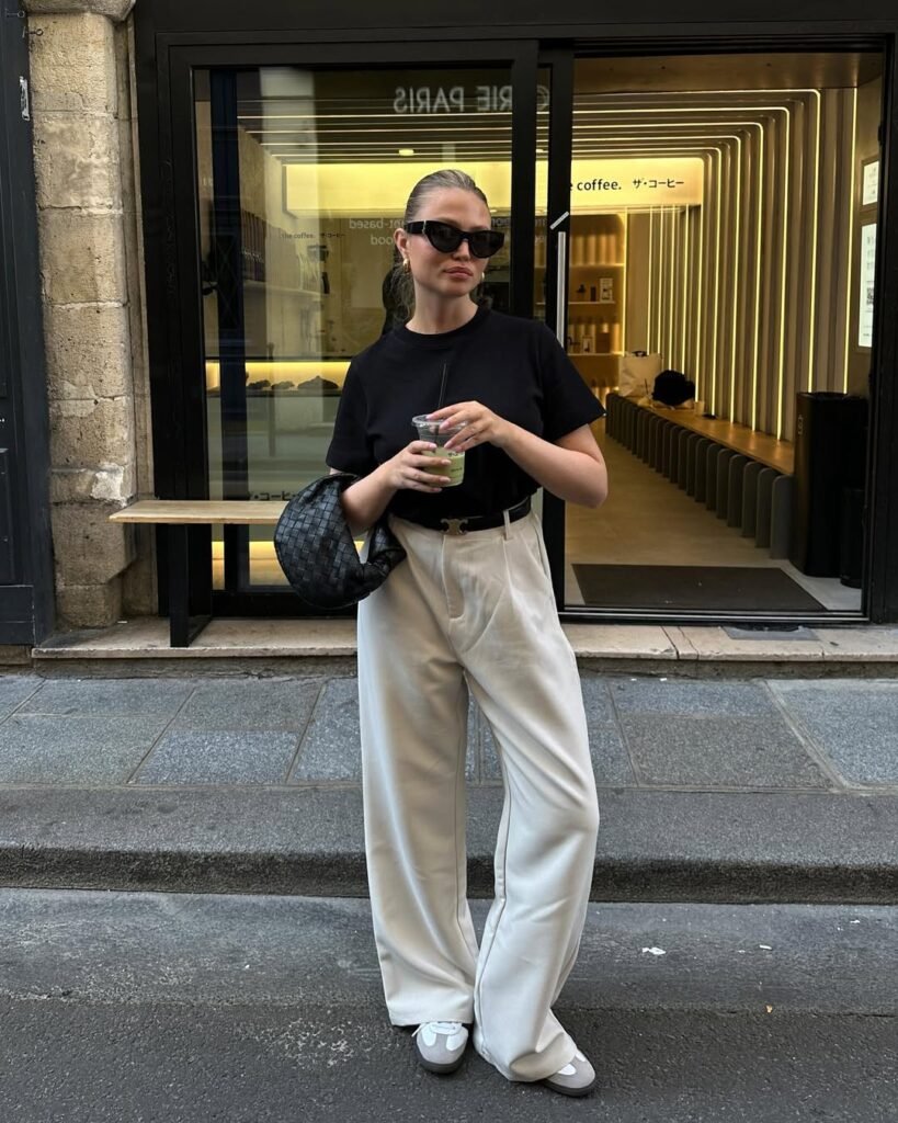 Woman wearing a black top and wide-leg cream trousers with ballet flats, holding a drink — a minimalist stylish work outfit for a casual office day.
