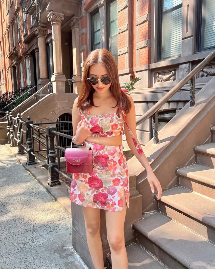 Young woman wearing a matching floral crop top and mini skirt set with a pink handbag and sunglasses, standing on a city staircase under bright sunlight — Outfits verano.