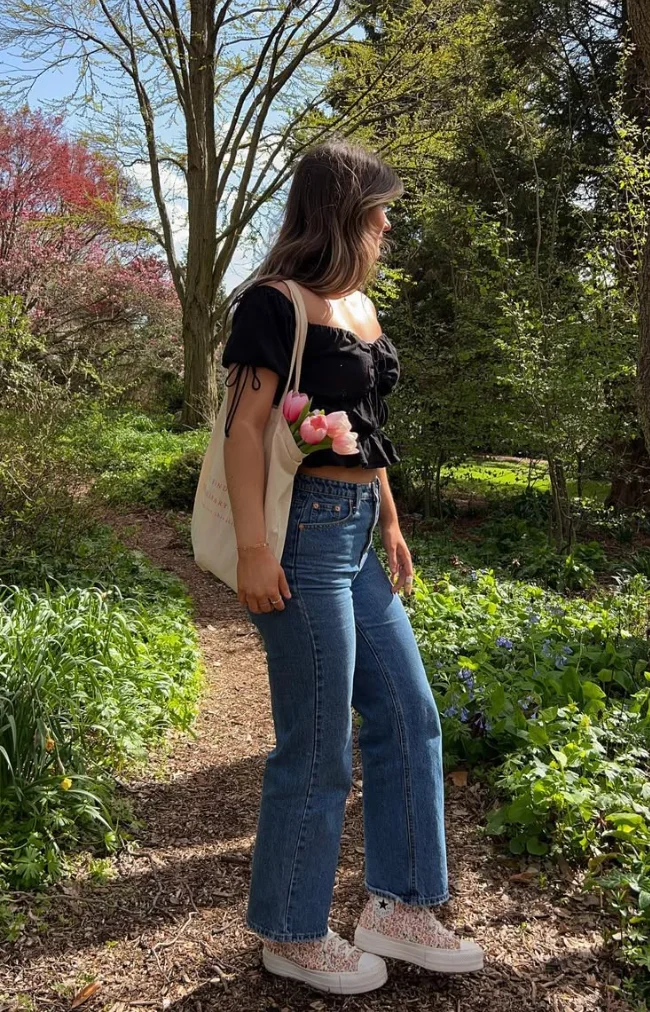 Picnic Date Outfits: Woman in a black ruched crop top and blue jeans walking through a wooded garden trail carrying tulips in a tote.