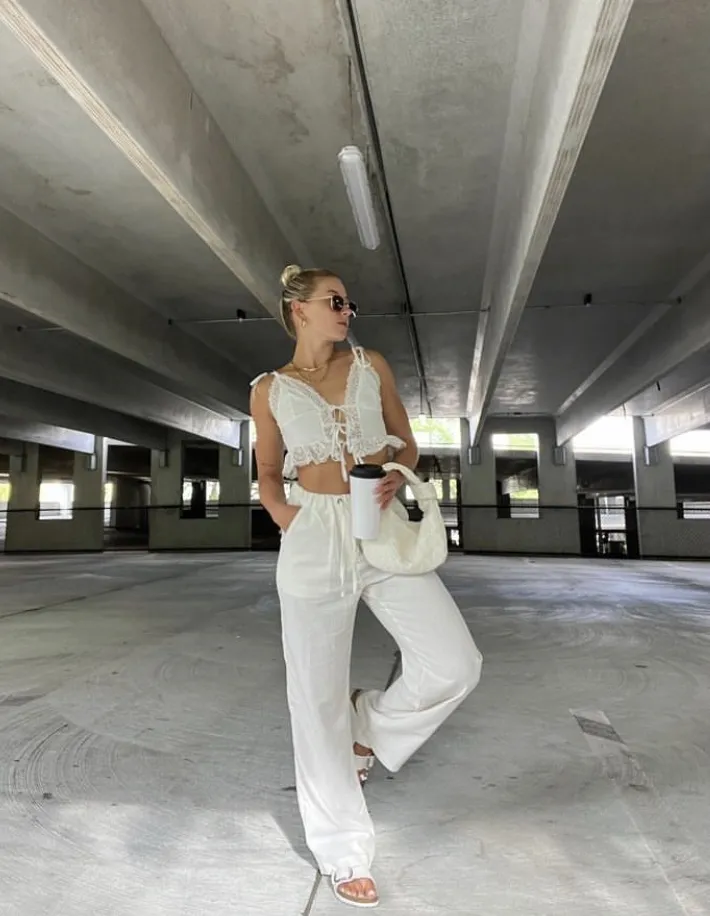 Blonde woman wearing loose white linen pants and a white lace crop top, posing in a parking garage with a white handbag and sandals. Casual linen pant outfit.