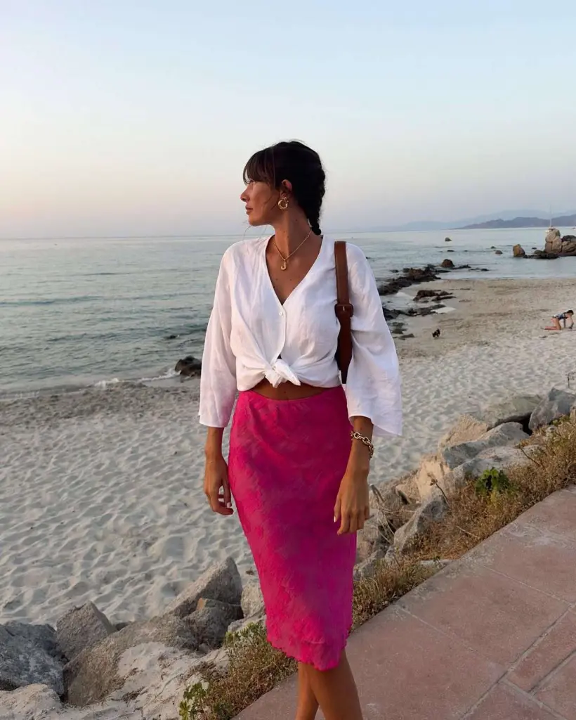 Beach outfit featuring a crisp white button-up shirt tied at the waist paired with a vibrant pink midi skirt, accessorized with gold jewelry and a brown shoulder bag by the seafront.