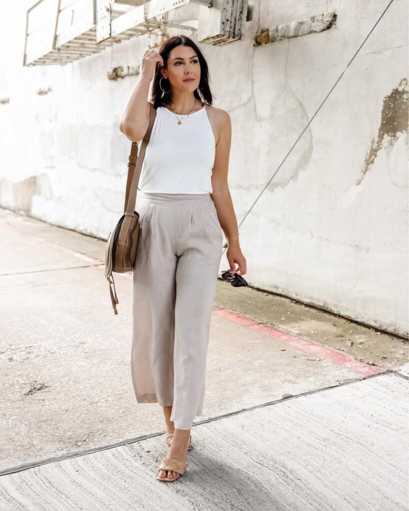 Woman walking outdoors in beige linen pants and a white tank top, accessorized with a tan crossbody bag and sandals. Simple and chic linen pant outfit.