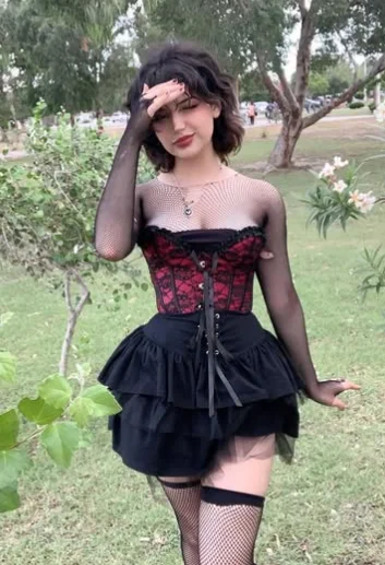 Grunge Outfits: Person with short dark hair wearing a black and red corset over a torn fishnet top, black layered mini skirt, and fishnet stockings, posing outdoors in a park.