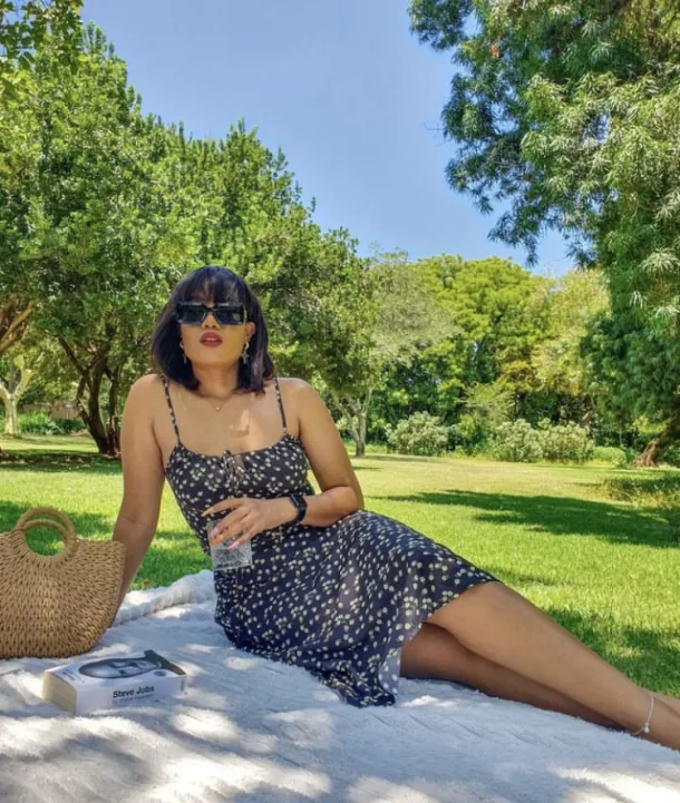 Picnic Date Outfits: Woman lounging on a blanket in a black and white polka dot dress with sunglasses, a woven bag, and a book.