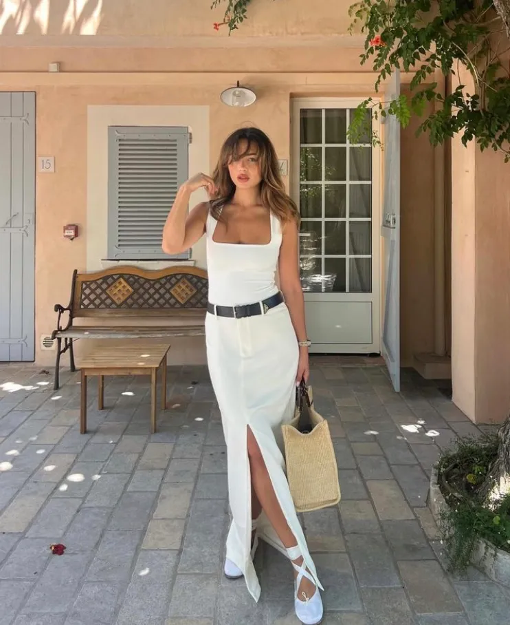A woman in an elegant white sleeveless top and high-slit skirt, styled with white flats and a woven tote bag — a polished and minimal European summer outfit.