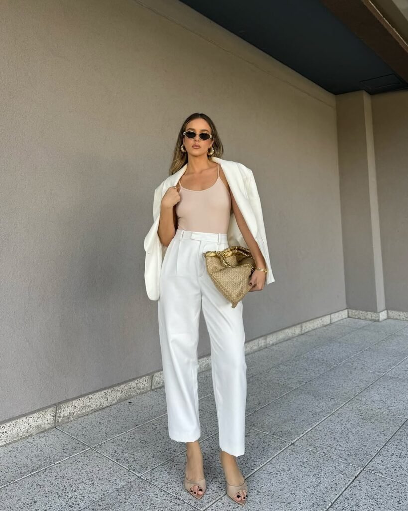  Elegant woman in a white blazer over a nude bodysuit and high-waisted white pants, accessorized with a straw clutch — a powerfully chic and stylish work outfit for summer meetings.