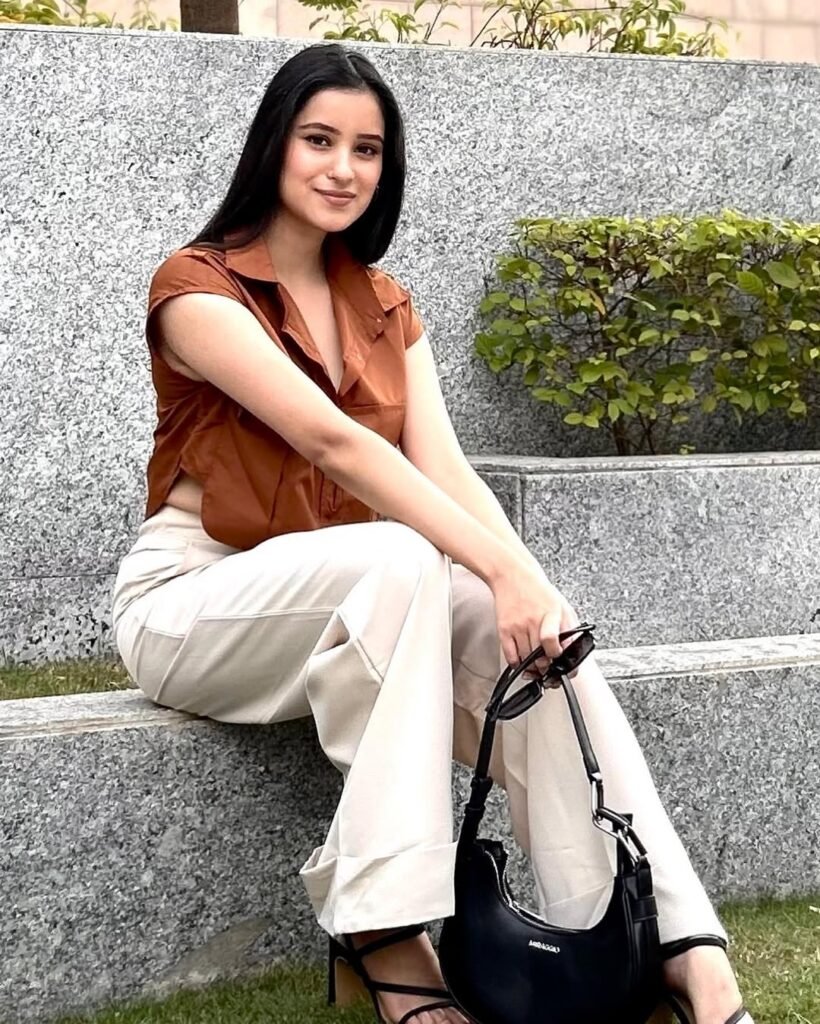  A woman dressed casually in a brown short-sleeve shirt and beige wide-legged trousers, sitting on a stone bench with a black handbag, exuding a sophisticated yet comfortable vibe suitable for a summer brunch date