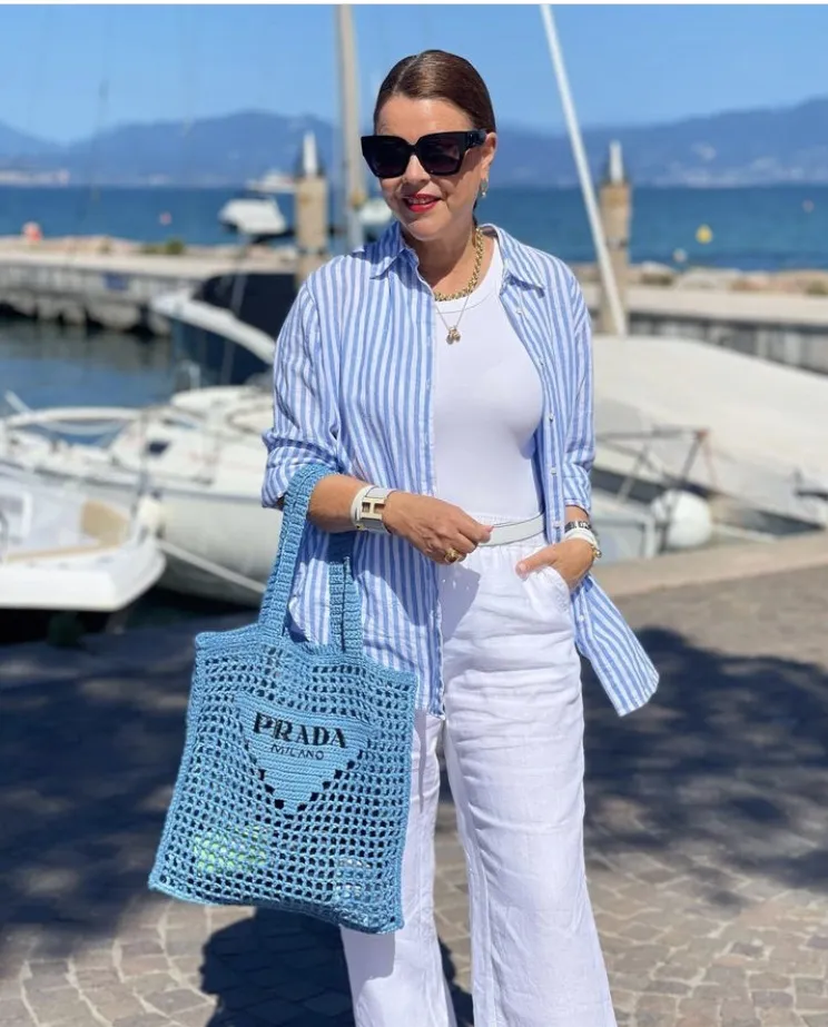 Woman by the harbor wearing white wide-leg linen pants with a blue striped shirt over a white tank, holding a blue Prada tote. Trendy linen pant outfit.