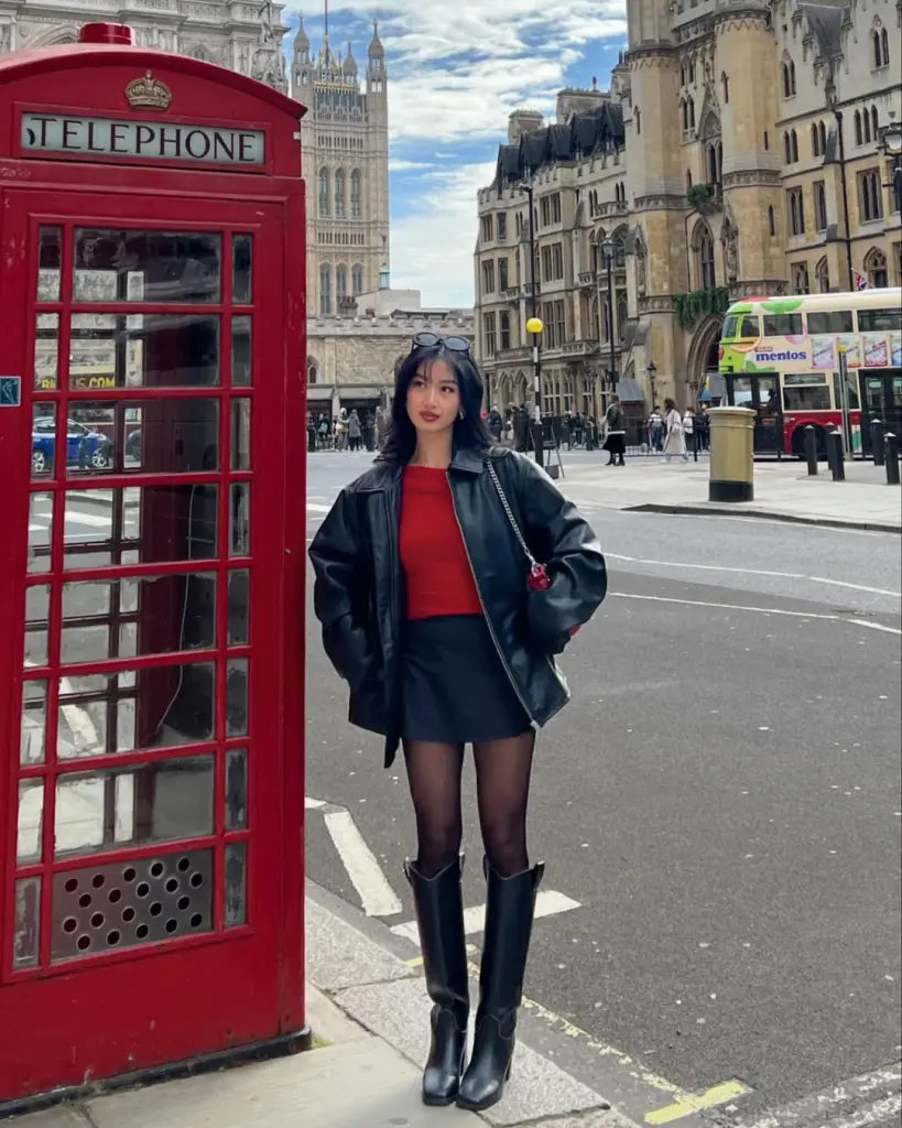 Edgy yet classy outfit with a red top, black mini skirt, sheer tights, and knee-high boots, layered under a bold black leather jacket, shot against iconic London architecture.