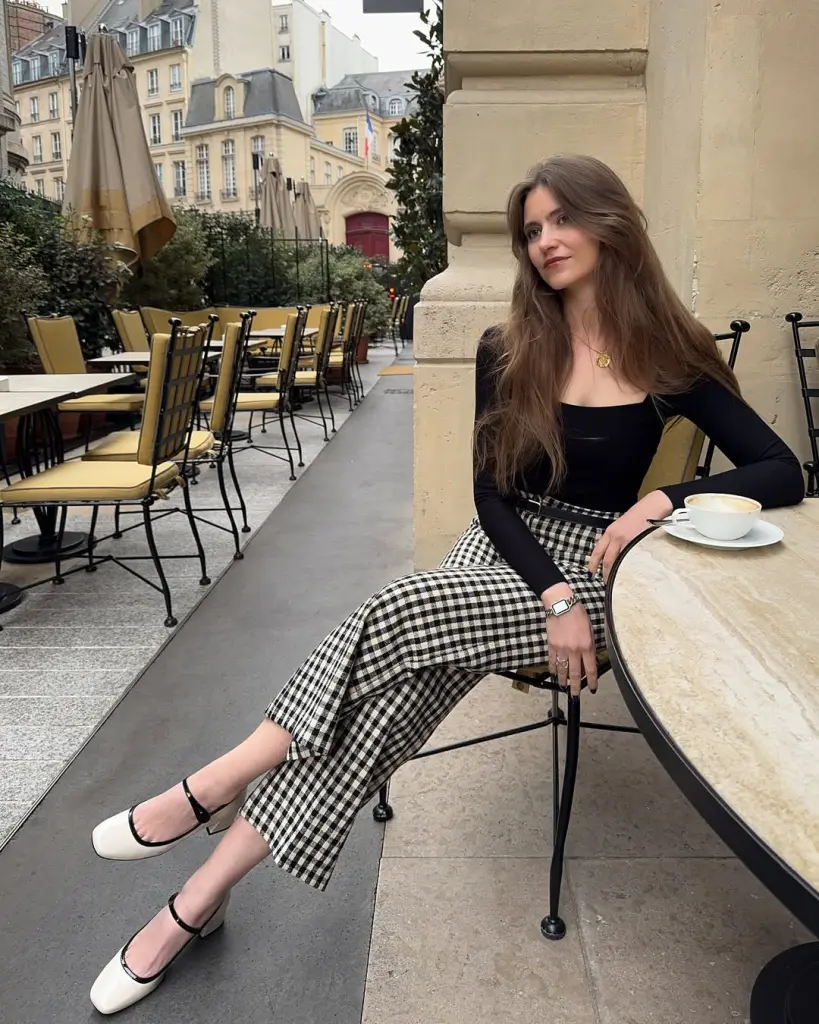 A woman seated at a Parisian café in a black square-neck top and checkered wide-leg pants, styled with white Mary Jane shoes — European summer style.

