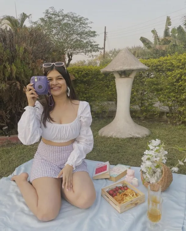 Picnic Date Outfits: Smiling woman in a white puff-sleeve crop top and purple shorts, holding a camera beside a colorful picnic spread.