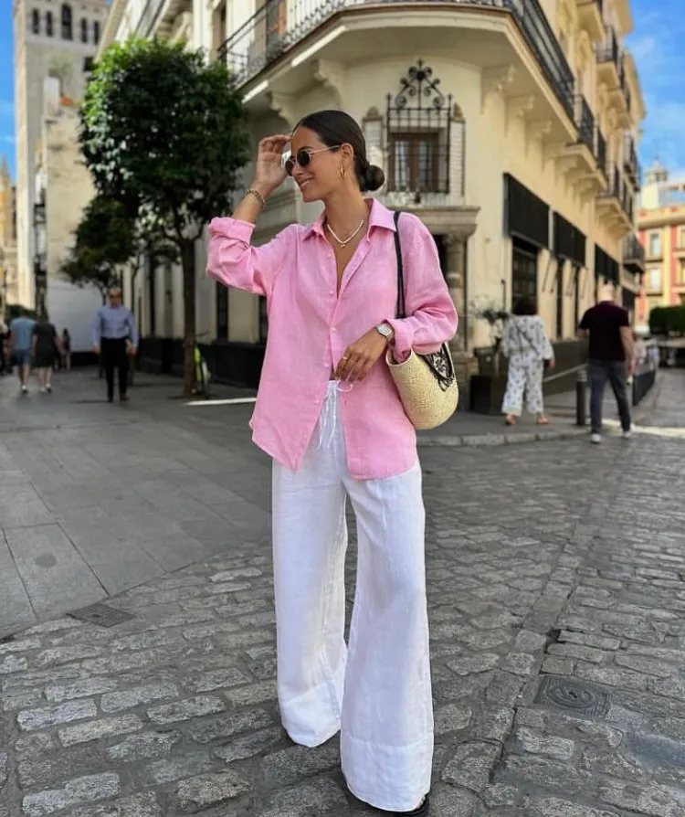 Woman in white linen pants paired with a pink oversized linen shirt, carrying a woven bag and wearing flat sandals. Casual and colorful linen pant outfit.