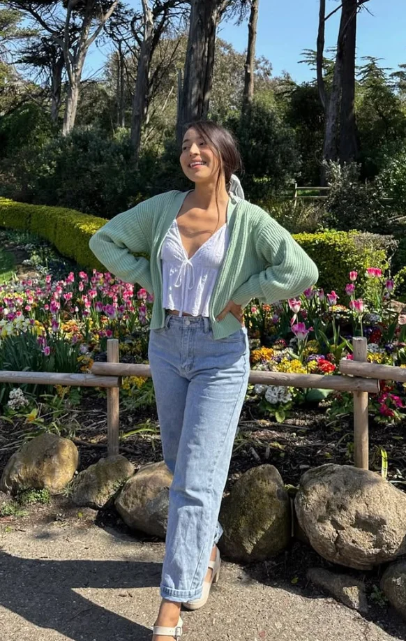 Picnic Date Outfits: Woman in a white top, mint cardigan, and blue jeans posing in a flower garden with a cheerful, spring vibe.