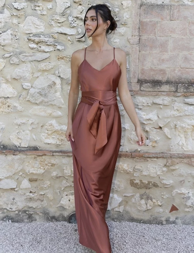 A woman in a satin rust-colored dress with thin straps and a tie belt, standing in front of a stone wall, giving an elegant look.